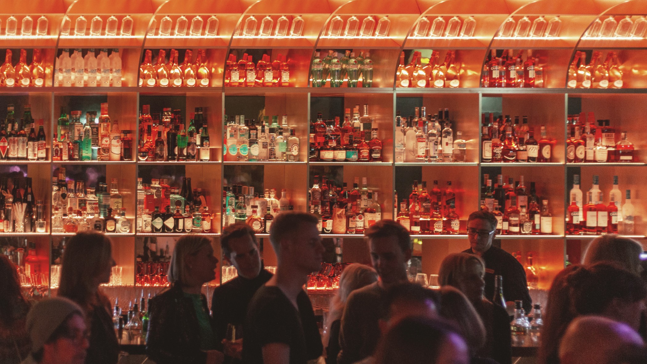 Gäste vor der hell erleuchteten Flaschenwand mit Rundbögen an der Bar