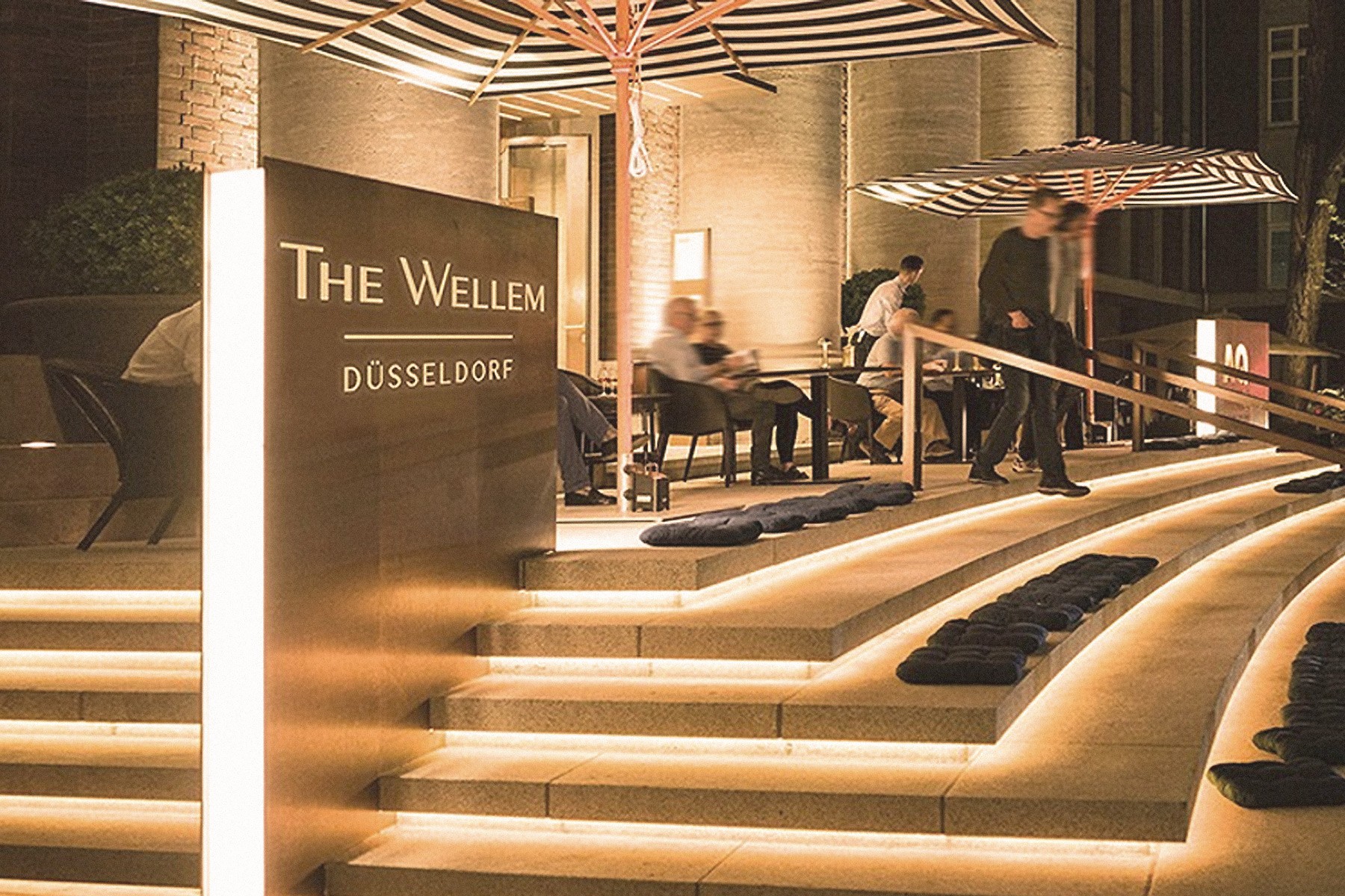 Beleuchtete Freitreppe mit Sitzkissen und Schild The Wellem Düsseldorf, Gäste auf der Terrasse am Abend