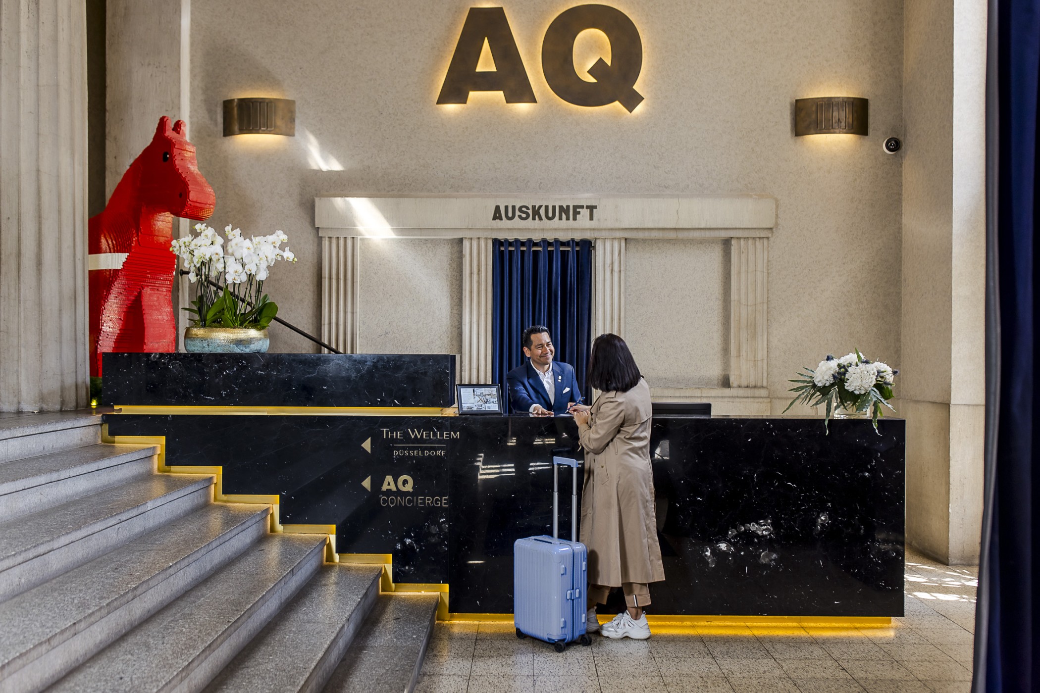 Concierge-Empfang von The Wellem mit AQ-Schriftzug und roter Pferdeskulptur, ein Gast mit Koffer am Tresen